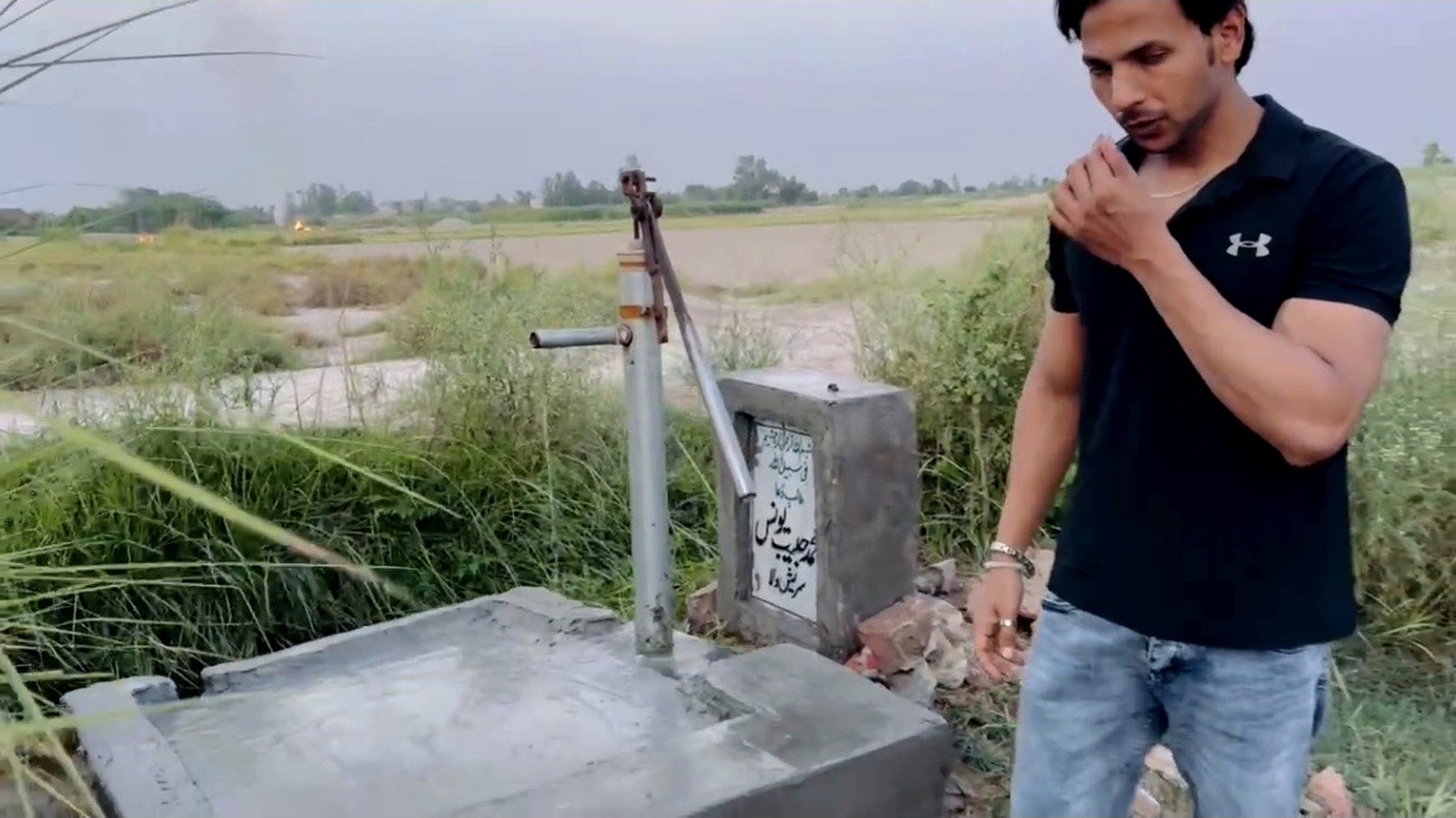 A hand pump installed on a concrete base beside farmland
