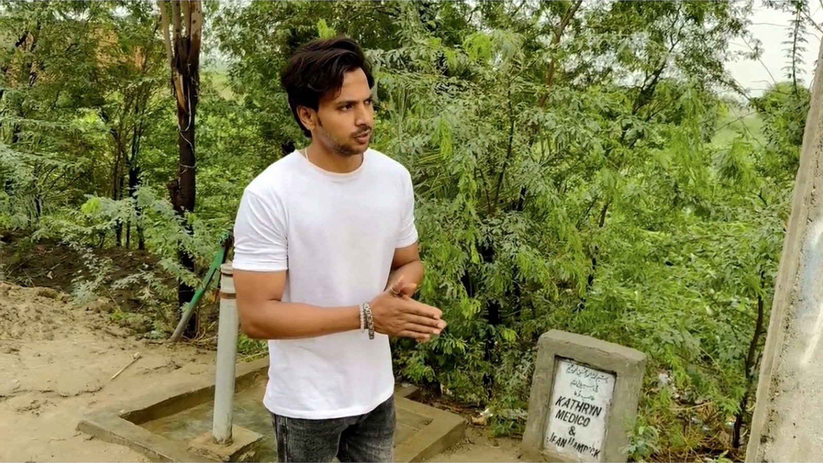 A man standing beside a hand pump surrounded by green trees
